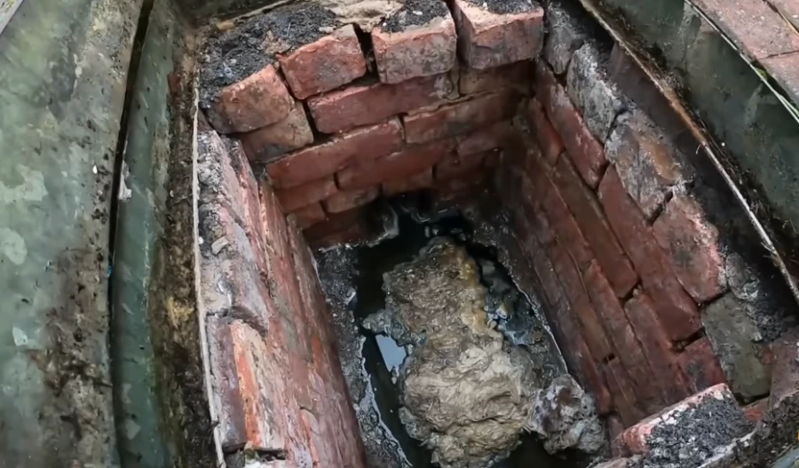 A brick-lined sewer chamber with a large grease and root blockage in standing water, illustrating aging pipe issues common near Comstock Park