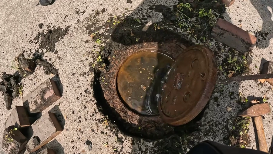 An old rusted metal cleanout cover pried open next to broken bricks, showing a hand-dug access point to an aging Spokane sewer line