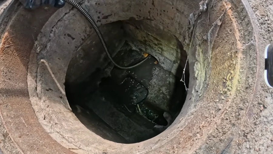 Looking down into a deep brick and concrete sewer manhole chamber with algae-stained walls and a cable descending into dark water, illustrating the structural condition of an aging Spokane sewer line